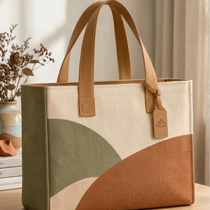 May include: A rectangular canvas tote bag featuring a color-blocked design in olive green, cream, and terracotta brown. The bag has two tan leather handles and a matching leather luggage tag embossed with a mountain icon hanging from one handle.