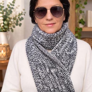 May include: A chunky knit scarf featuring a marled pattern of black, white, and grey yarn. The scarf is wrapped around the neck in a thick, ribbed texture. The accessory is paired with dark-tinted aviator sunglasses with thin gold-tone frames.