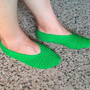 May include: A pair of bright green knitted slippers featuring a textured, woven stitch pattern. The slippers have a low-cut, slip-on design that covers the toes and sides of the feet, leaving the top of the foot exposed in a simple, rounded opening.