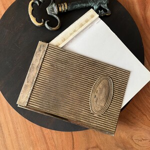 Può includere: Un astuccio metallico rettangolare con texture a coste e una placca ovale liscia sul davanti, dotato di un lato incernierato. Un cartoncino rettangolare bianco è parzialmente inserito nella parte superiore. Una chiave antica in bronzo decorata si trova accanto.
