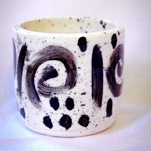 May include: A cylindrical ceramic cup with an off-white base featuring black speckled dots and large, hand-painted black brushstroke swirls. The design includes vertical black lines and circular patterns wrapping around the exterior surface.