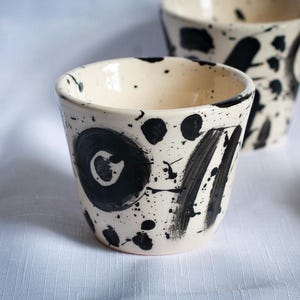 May include: Two ceramic cups with a cream-coloured base featuring hand-painted black abstract designs. The exterior of each cup displays bold black circular motifs, thick brush strokes, and scattered black paint splatters across the surface.