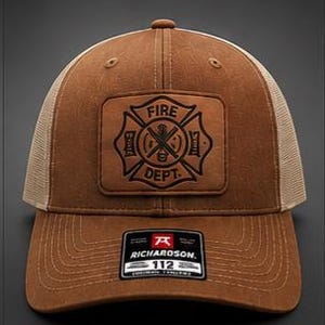 May include: A brown and khaki trucker hat featuring a rectangular leather patch on the front with a black embossed Fire Dept Maltese cross emblem. A black sticker on the brim displays the Richardson 112 logo and text. The hat has mesh back panels.