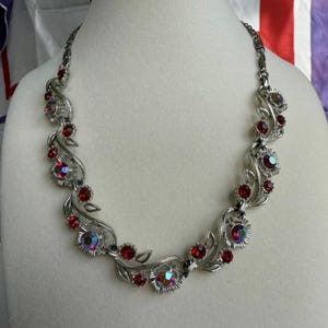 Puede incluir: Un collar de metal en tono plateado con un diseño de enredadera y hojas pulidas. La pieza está adornada con pequeñas piedras redondas de color rojo intenso facetadas y cristales circulares iridiscentes y multicolores en la cadena.