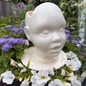 Puede incluir: Una maceta de cerámica blanca brillante con forma de cabeza humana y parte superior hueca. La escultura presenta un cráneo liso y redondeado, rasgos faciales definidos que incluyen ojos, nariz y labios entreabiertos, sobre una base circular.