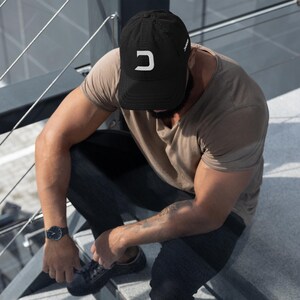 Puede incluir: Una gorra de béisbol negra con un logotipo de la letra D en blanco en el panel frontal. La gorra tiene una visera curva y una corona estructurada. El diseño es sencillo y funcional, destacando el contraste entre la tela negra y la letra.