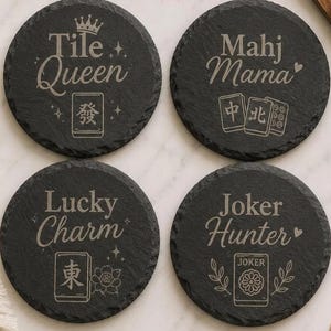 May include: Four round black slate coasters with rough edges, each engraved with unique Mahjong-themed designs and text. The coasters feature the phrases Tile Queen, Mahj Mama, Lucky Charm, and Joker Hunter, accompanied by various Mahjong tile illustrations.