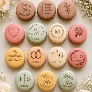 May include: A collection of fourteen round macarons in various pastel colors, each featuring custom laser-engraved designs including text like Mr. & Mrs., T & G, dates, floral motifs, heart shapes, and wedding ring illustrations on their smooth surfaces.