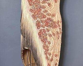 May include: A beige asymmetrical dress featuring a one-shoulder neckline and a short sleeve. The garment is adorned with intricate copper and pink beaded floral patterns and long, flowing fringe that transitions from beige to dark brown at the hem.