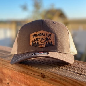 May include: A brown and beige trucker hat with a brown leather patch that says "Grandpa Life" with a mountain range and trees design.