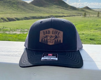 Pode incluir: Um boné de caminhoneiro preto e cinza com um remendo de couro marrom que diz "Dad Life" com uma cena de montanha. O boné é feito pela Richardson 112.