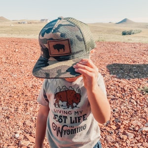 May include: A child wearing a camouflage trucker hat with a brown leather patch featuring a silhouette of a buffalo. The hat has a mesh back. The child is also wearing a light gray t-shirt with a brown buffalo graphic and the text "Living my Best Life in Wyoming".