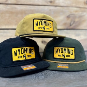 Könnte beinhalten: Drei Baseballkappen mit gelben und schwarzen Wyoming-Patches. Die Patches haben den Text "Wyoming Est. 1890" und eine Silhouette eines Cowboys, der ein Pferd reitet. Die Kappen sind in verschiedenen Farben: Khaki, Schwarz und Grün.