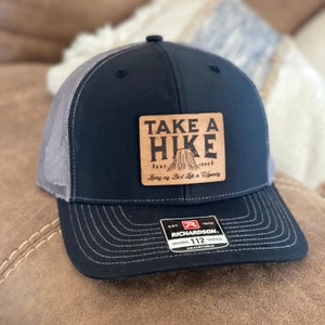May include: A black and gray trucker hat with a brown leather patch that says "Take a Hike" with a mountain silhouette and the text "Loving my Best Life in Wyoming". The hat has a Richardson logo and the text "Original 112 Trucker Adjustable".