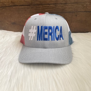 May include: A gray trucker hat with a red, white, and blue American flag design on the back. The front of the hat has the text "#MERICA" embroidered in blue.