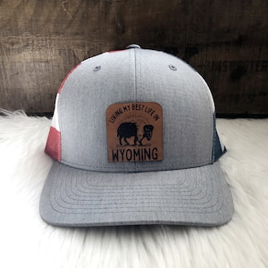 May include: A gray trucker hat with a red, white, and blue American flag design on the side. The hat has a brown leather patch on the front that says "Living my best life in Wyoming" with a silhouette of a buffalo.