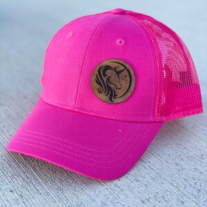 May include: Pink trucker hat with a brown leather patch featuring a unicorn silhouette. The patch has a moon and stars design.