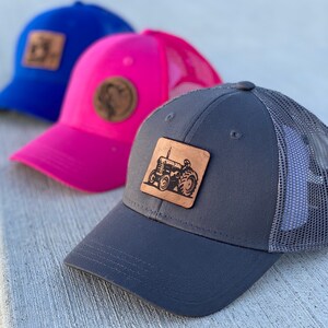 May include: Three trucker hats with different colored brims and mesh backs. The hats have leather patches with a tractor design. The hats are blue, pink, and gray.