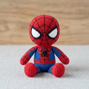 May include: A crocheted doll of a superhero character with a round head, large white eyes, and a red and blue suit. The red sections feature a black web pattern, and a small black spider emblem is centered on the chest. The doll is in a seated position.