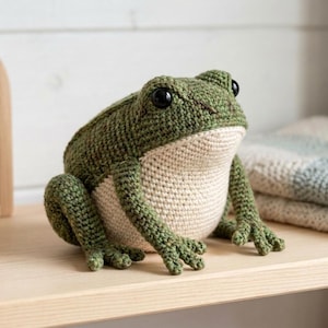 May include: A handcrafted crochet frog toy featuring a textured dark green back and a smooth cream-colored belly. The plush figure has large, round, glossy black eyes and detailed webbed feet, sitting in a natural, upright, and alert posture.