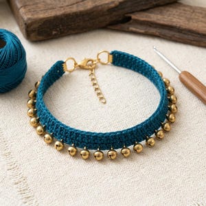 May include: A teal crocheted choker necklace featuring a series of small, round, metallic gold-toned bells attached along the bottom edge. The necklace includes a gold-toned lobster claw clasp and an adjustable chain extender for a secure fit.