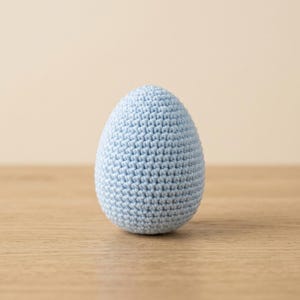 Puede incluir: Un objeto con forma de huevo de color azul claro elaborado con un patrón de punto de ganchillo denso y uniforme. El artículo presenta una parte superior redondeada y una base ligeramente más ancha, con una textura consistente en azul.