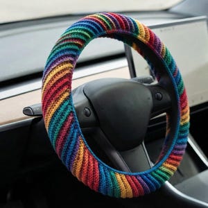 May include: A circular steering wheel cover featuring a vibrant, multi-colored crochet pattern with diagonal stripes in red, orange, yellow, green, blue, and purple. The textured, ribbed yarn construction fits snugly around the black steering wheel.