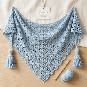 May include: A light blue hand-crocheted lace shawl with a triangular shape, featuring a repeating fan pattern and two matching tassels at the corners. A white tag reads WINTER ICE Hand Crochet Lace Shawl. A wooden crochet hook and yarn are included.