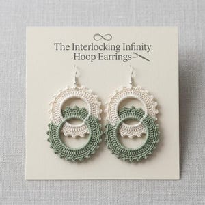 May include: A pair of crochet hoop earrings featuring two interlocking circles in cream and sage green thread. The earrings are displayed on a cream-colored card with the text The Interlocking Infinity Hoop Earrings printed in dark gray.