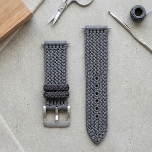 Op de afbeelding: Een grijze gevlochten horlogeband met een geweven koordtextuur, twee bijpassende lussen en een zilverkleurige metalen gesp. De band bevat een reeks verstevigde ronde gaten voor aanpassing, uitgevoerd in een gedetailleerd en ingewikkeld knoppatroon.
