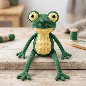 Puede incluir: Un juguete de rana hecho a ganchillo con un cuerpo verde oscuro, vientre amarillo claro y ojos grandes de color amarillo pálido con pupilas negras. La rana tiene brazos y piernas largos y delgados con pies de varios dedos hechos de hilo.