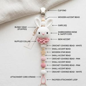 May include: A pacifier clip featuring a stuffed white crochet bunny head with embroidered nose and safety eyes, a pink ruffled collar, and a bow accent. The strand includes white and pink crochet-covered beads, wooden accent beads, and a wooden star bead, ending in a pink attachment loop and a metal clip.