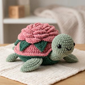 Pode incluir: Um peluche de tartaruga em croché com cabeça e membros verde-claros, barriga verde-escura e uma carapaça desenhada como uma grande flor de rosa rosa texturizada com folhas verde-escuras a rodear a base das pétalas da flor.