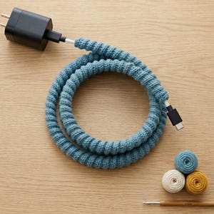 May include: A black charging cable encased in a textured, blue knitted yarn sleeve, coiled into a circle. Beside the cable are three small, round balls of yarn in blue, white, and yellow, along with a wooden crochet hook marked 3.5mm.