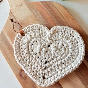 May include: A cream-colored, heart-shaped crochet trivet with a leather loop for hanging. The trivet is placed on a wooden cutting board. The trivet is made of thick, textured yarn. The wooden cutting board has a light brown color.