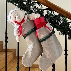 Pode incluir: Duas meias de Natal de linho bege com acabamento em renda verde-escura no topo. Uma meia contém um urso de peluche creme com uma fita vermelha, a outra uma pequena caixa de presente vermelha atada com uma fita de cetim vermelha.