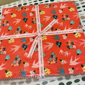 以下が含まれることがあります： 鮮やかなコーラル色の生地の正方形の束。青、黄色、白の色合いの花と鳥の模様で飾られています。「Poppie Cotton sunshine and joy in every detail」と書かれたリボンで束ねられています。