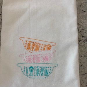 May include: A white kitchen towel with three embroidered bowls stacked on top of each other. The bowls are decorated with a folk art design in orange, pink, and teal.