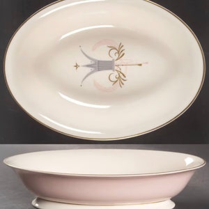 May include: Oval serving dish with a cream interior and a pink exterior. It has a gold rim and a central design in grey, gold, and pink. The dish is raised on a white base. A classic piece of tableware.