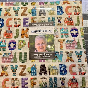Könnte beinhalten: Ein Stoffquadrat mit einem sich wiederholenden Muster aus bunten Buchstaben und kleinen Hausillustrationen. Die Buchstaben sind in verschiedenen Schriftarten und -stilen gehalten, mit Vintage-inspirierten Designs. Ein Etikett mit einem Frauenporträt und dem Text "moda fabrics + supplies" befindet sich oben.