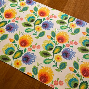 Puede incluir: Un camino de mesa blanco con un estampado floral colorido. Las flores son rojas, amarillas, naranjas, azules, verdes y moradas. El camino de mesa tiene un fondo blanco con un sutil estampado floral.