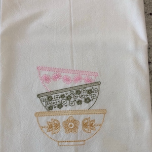 May include: A white kitchen towel with embroidered bowls in pink, green, and gold. The bowls are stacked on top of each other.
