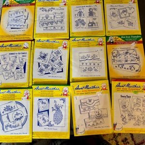 May include: A selection of vintage Aunt Martha's Hot Iron Transfer packets. The packets are yellow with designs in blue and white, featuring floral patterns, bunnies, and a peacock. These packets are for embroidery and quilting projects.