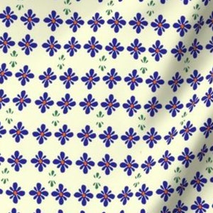 Op de afbeelding: Crèmekleurige stof met een herhalend patroon van blauwe bloemen met oranje centra. De bloemen zijn in rijen gerangschikt, met kleine groene bladachtige vormen tussen de rijen. De stof heeft een lichte textuur.