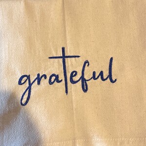 May include: A white kitchen towel with the word "grateful" embroidered in blue with a cross above the word.