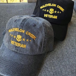 Könnte beinhalten: Zwei Baseballkappen, eine schwarze und eine verwaschene blaue, mit goldfarbener Stickerei "MALEVELON CREEK VETERAN". Das Design beinhaltet einen Totenkopf und Sterne.
