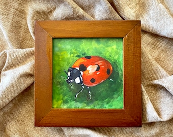 May include: A framed painting of a ladybug on a green leaf. The ladybug is red with black spots and white markings. The frame is brown wood.