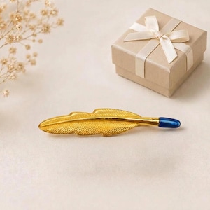 May include: A gold-toned metal hair clip shaped like a feather with a smooth, dark blue rounded tip. The feather design features fine, etched lines along its surface to mimic the texture of a quill, presented alongside a square gift box.