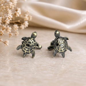 May include: A pair of cufflinks shaped like sea turtles, featuring a dark metallic finish with gold-toned accents on the shell patterns. The turtle designs are detailed with a segmented shell structure and extended flippers, presented as matching accessories.