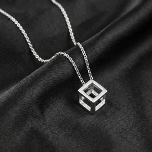 May include: A silver-coloured necklace featuring a pendant shaped like an open-frame cube. The pendant is suspended from a delicate, matching silver-coloured chain with small, uniform links. The design consists of thin, rigid metal bars forming a hollow cube.